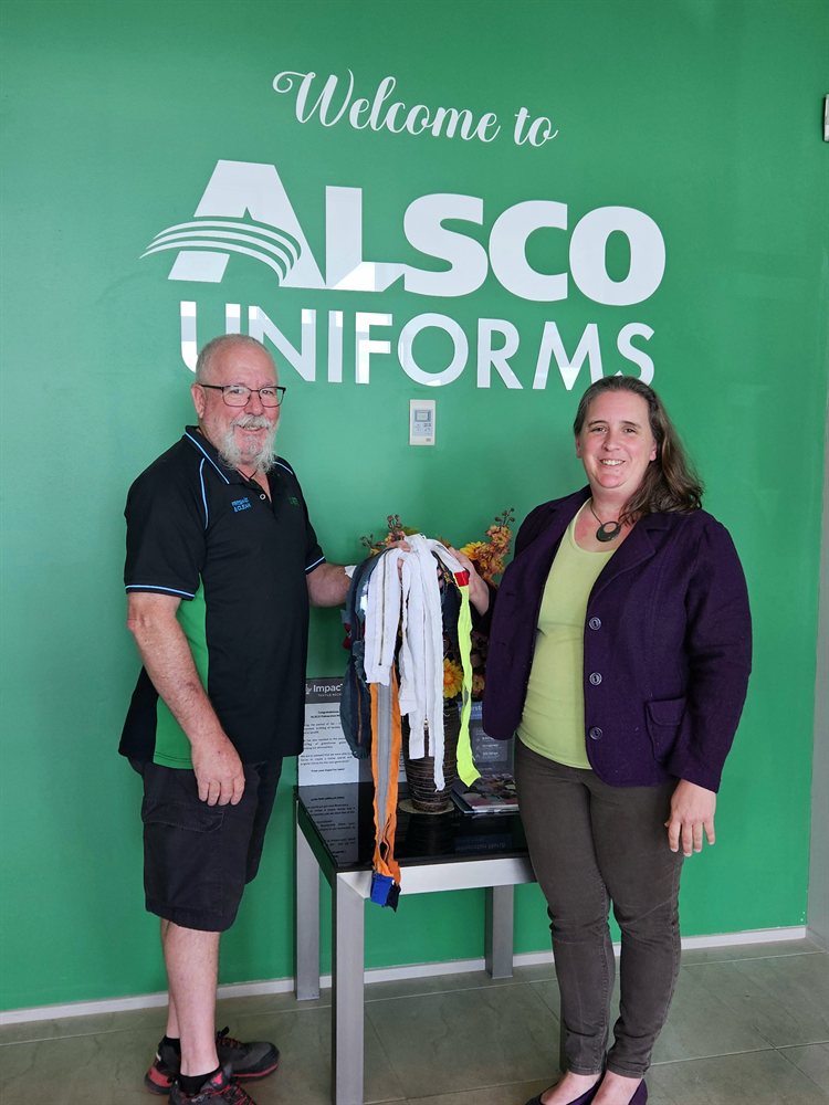 Two people with recovered zips and bed sheets in front of a 'Welcome to Alsco Uniforms' banner